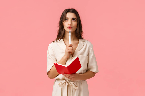 Thoughtful and creative young woman making list, pouting and looking up pensive and inspired, holding red notebook and pen, create new poem or prepare for exam, thinking over pink background - Foto, imagen