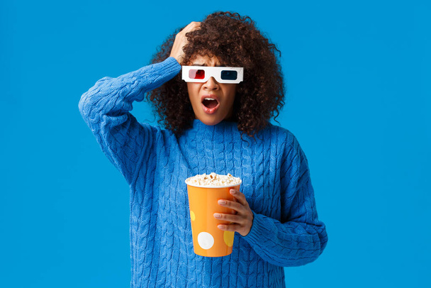 Shocked african-american pretty woman concerned with shocking cliffhanger in movie, grab head uneasy and upset, drop jaw, holding popcorn watching film in cinema, blue background - Photo, Image