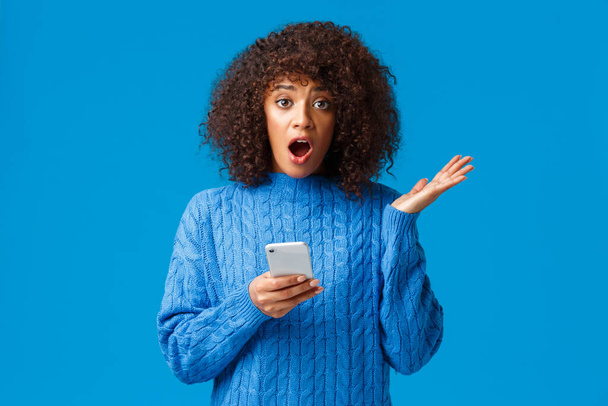 Bezorgd en bezorgd geschokt jonge Afrikaans-Amerikaanse vrouw ontvangen onaangenaam nieuws via smartphone, vertellen met besluiteloze blik, shrugging en raise hand dont know what do, blauwe achtergrond - Foto, afbeelding