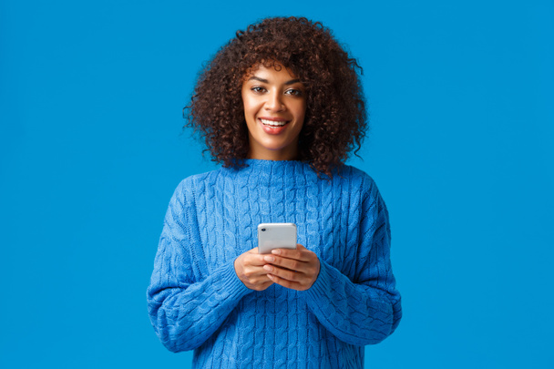 Addiction au smartphone, concept gen-z et personnes. Charismatique belle femme afro-américaine souriante avec coupe de cheveux afro en pull d'hiver, tenant smartphone et appareil photo, fond bleu - Photo, image