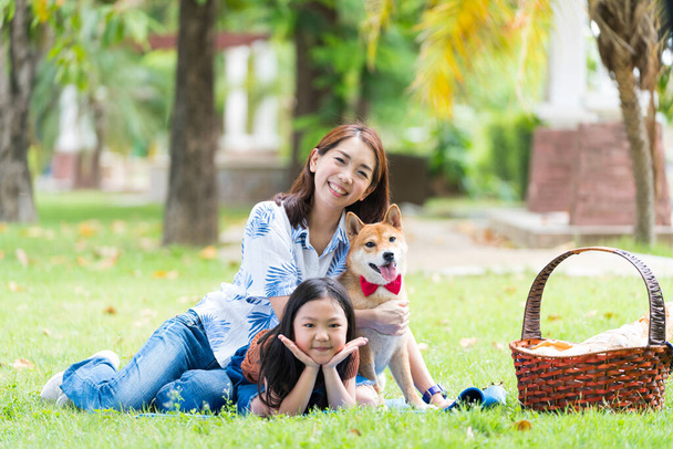 春に芝犬と一緒に公園に座っている母親と娘の家族。アジアの家族は庭でピクニックをしています。カメラを見ている女性は. - 写真・画像
