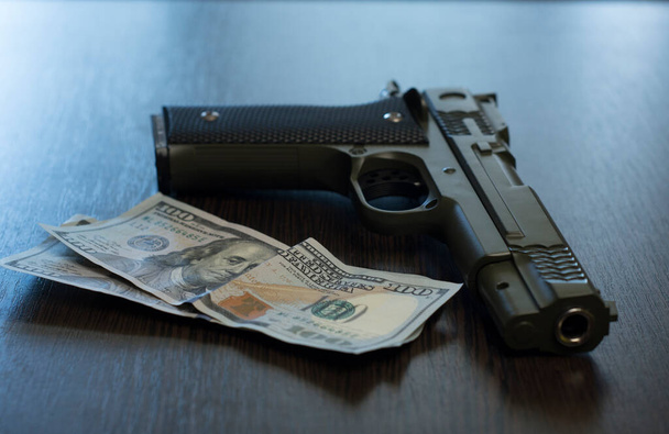Khaki pistol and two hundred dollar bills lie on a wooden table. Close up criminal concept - Photo, Image