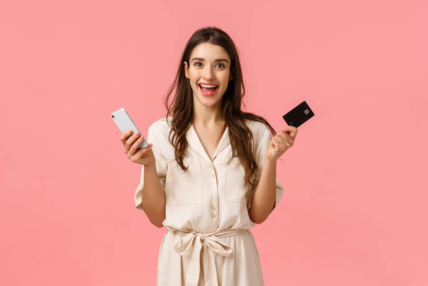 Winkelen, online betalen, lifestyle concept. Gelukkig verleidelijk meisje in jurk, met creditcard en smartphone, vrolijk glimlachen in afwachting van nieuwe product levering van internet winkel, roze achtergrond - Foto, afbeelding