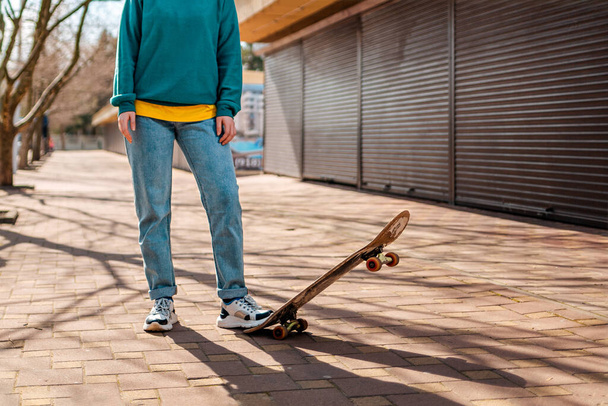 Подросток-хипстер позирует ногой на скейтборде и смотрит вдаль. На заднем плане переулок. Закрывай. Концепция спортивного образа жизни и уличной культуры
. - Фото, изображение