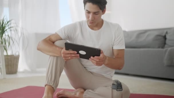 Smiling Young Man Surfing Internet on Tablet While Sitting on Exercise Mat During Relaxing After Workout at Home. - Imágenes, Vídeo
