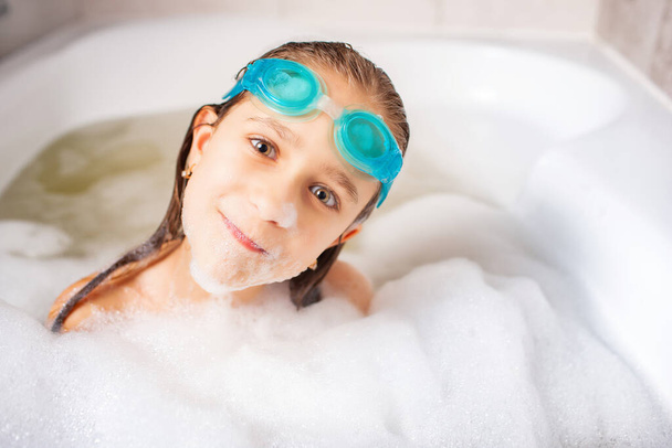 Funny little positive caucasian girl wearing swimming goggles and plays in the bath with foam while waiting for relaxation by the sea. Concept of hygiene and home entertainment for children - Valokuva, kuva