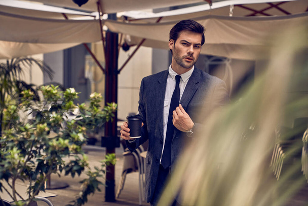 Pauza na kafe. Sebevědomý mladý muž v plném obleku drží šálek kávy a dívá se stranou, zatímco stojí venku s městskou krajinou v pozadí - Fotografie, Obrázek