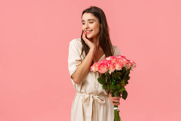 Нежность, женщины и концепция красоты. Привлекательная чувственная молодая девушка получает красивые букеты цветов, держа розы, глядя в сторону с застенчивой милой улыбкой, стоя розовый фон
 - Фото, изображение