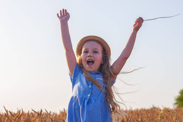 Urocza dziewczynka w słomkowym kapeluszu i niebieskiej sukience w kratę na polu pszenicy z rękami do góry, uśmiechnięta do nieba. Szczęśliwe dziecko z długimi blond falowane włosy krajobraz wsi. Zabawny spacerowicz na świeżym powietrzu - Zdjęcie, obraz