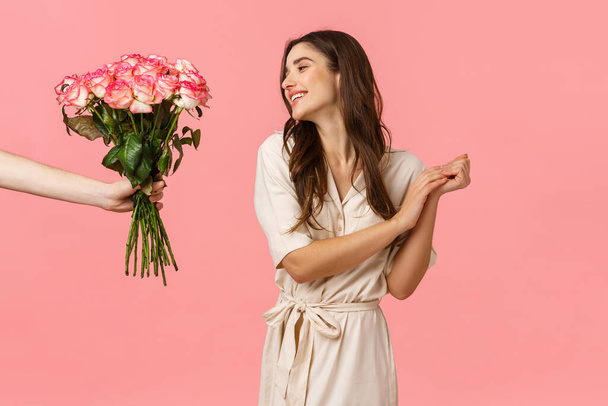 Romans, walentynki, koncepcja miłości. Urocza młoda kobieta wzdychająca romantycznie i zachwycona, patrząc na dłoń dając kwiaty, piękne bukiet róż, uczucie pielęgnacji, różowe tło - Zdjęcie, obraz