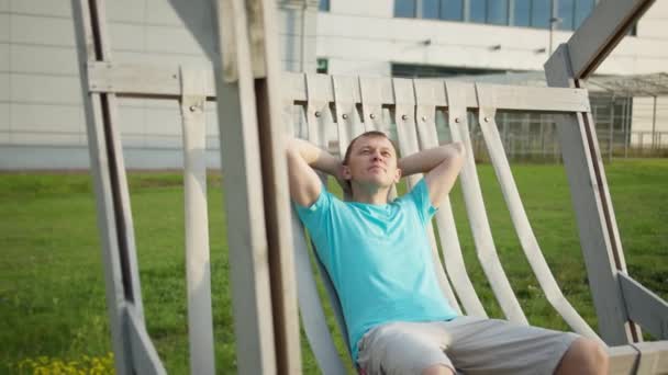 Mężczyzna odpoczywa w parku z rękami za głową, siedzi na wiklinowej ławce, rusza kamerą - Materiał filmowy, wideo