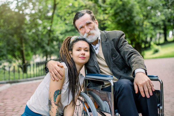 Female young social worker and disabled man on a walk. Girl and grandfather sitting in a wheelchair, hugging and posing to camera, while walking in the park. Concept of help for the elderly - Φωτογραφία, εικόνα