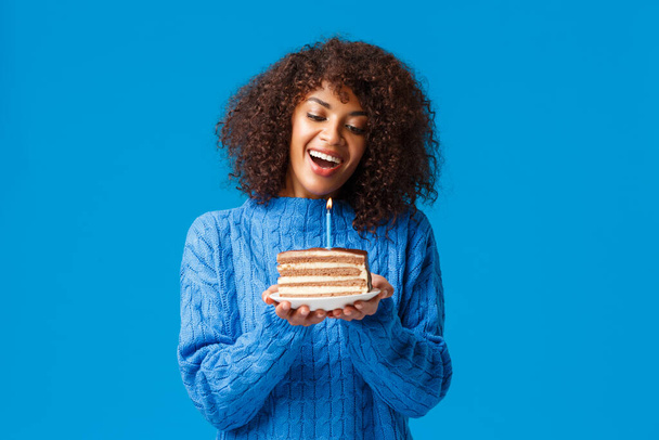 Celebração, feriados e conceito de festa. Sonhador e linda mulher afro-americana bonito com corte de cabelo afro, em suéter, inclinar a cabeça e assistir a vela acesa no bolo de aniversário, sorrindo fazendo desejo
 - Foto, Imagem