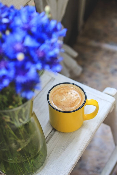 na varanda é uma mesa de madeira branca com uma xícara de café amarelo e um vaso com flores de milho. - Foto, Imagem