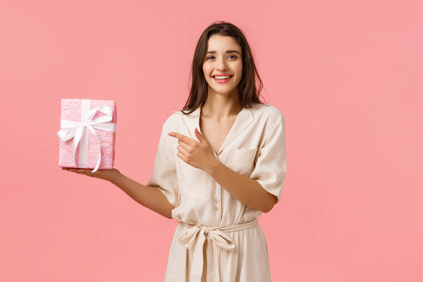 Girl prepared romantic gift for anniversary. Cheerful pretty young woman in cute dress holding gift box and pointing finger at present with delighted, happy smile, standing pink background - Фото, изображение