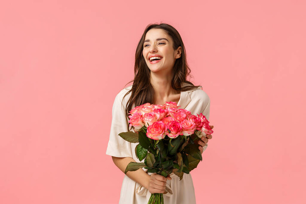 Valentines day, romance, celebration and beauty concept. Charming elegant and feminine young woman receive flowers, holding beautiful bouquet and laughing joyfully, pink background - Photo, image