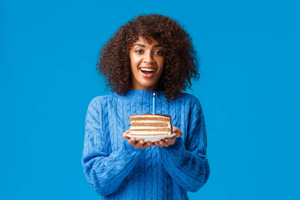 Make a wish and it come true. Cheerful and dreamy cute african-american b-day girl, holding cake with candle, blowing-out and smiling, having birthday party, standing in sweater blue background - Foto, Bild