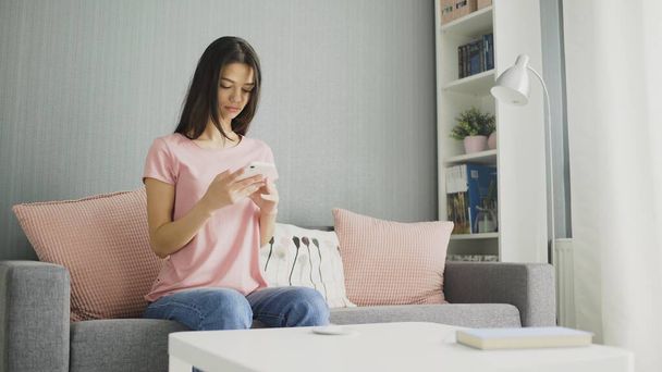 Mladá sexy brunetka žena pomocí telefonu, zatímco sedí na gauči doma. - Fotografie, Obrázek