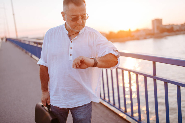 Белый бизнесмен средних лет в очках, в белой рубашке и с черным портфелем в руке идет по мосту и смотрит на часы
 - Фото, изображение