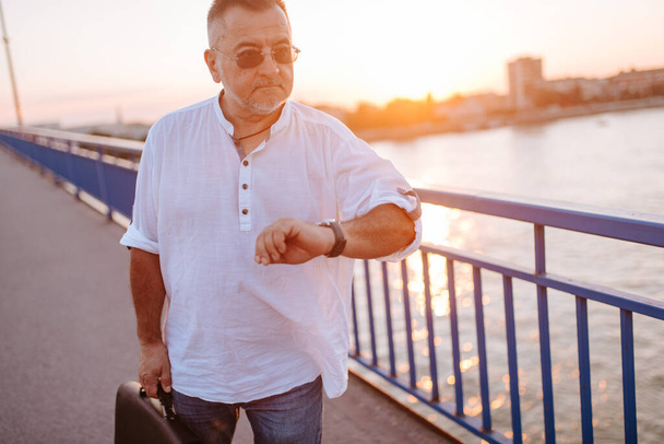 Un homme d'affaires caucasien d'âge moyen avec des lunettes, une chemise blanche et une mallette noire à la main marche sur le pont et regarde sa montre - Photo, image