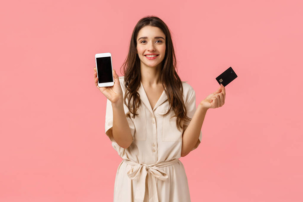 Δώρα, ψώνια και ομορφιά. Μοντέρνα νεαρή γυναίκα σε μοντέρνο φόρεμα, κρατώντας smartphone και πιστωτική κάρτα, χαμογελώντας χαρούμενα, να παραγγείλετε το προϊόν σε απευθείας σύνδεση, στέκεται ροζ φόντο - Φωτογραφία, εικόνα