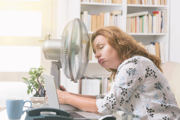 Женщина страдает от жары во время работы в офисе и пытается остыть за счет вентилятора
 - Фото, изображение