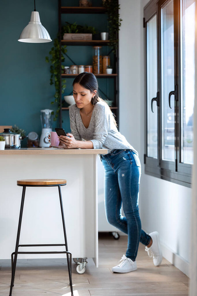 Shot of concentrated young woman using her mobile phone while drinking a cup of coffee in the kitchen at home. - Valokuva, kuva