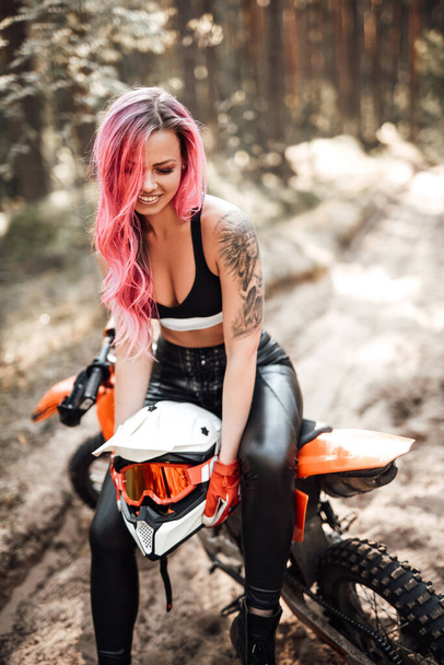 Молодая гонщица с розовыми волосами и татуировкой под рукой в спортивном лифчике сидит на мотоцикле - Фото, изображение