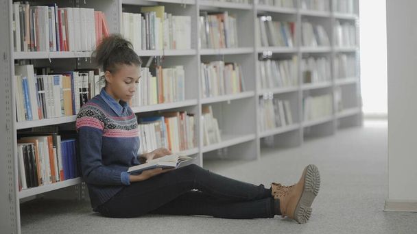 Young african american woman is reading book sitting on floor in university library. - Фото, зображення