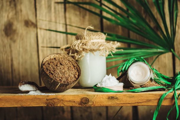 Kokosöl mit frischer Nuss auf altem Holztisch. Kokosöl und Kokosnüsse. Bio-Kokosnussprodukte für den Wellnessbereich. Natürliche reinigende Wellness-Accessoires und Peeling-Badezimmerprodukte auf Holz-Hintergrund. - Foto, Bild
