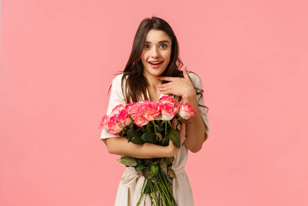Surprised girl astonished and touched with beautiful roses boyfriend brought on date. Attractive young woman in dress, smiling and gasping flattered, holding stunning bouquet, pink background - Foto, Bild