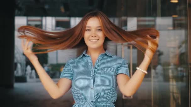 Schöne lächelnde glückliche Mädchen in gestreiftem Kleid reißt anmutig lange Haare gegen die Spiegelwand eines modernen Restaurants an einem Sommertag, Nahaufnahme in Zeitlupe. Positive junge Frau. - Filmmaterial, Video