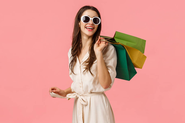 Winkelen, levensstijl en consumentenconcept. Aantrekkelijke jonge zorgeloze meisje in bril en jurk, kijk achter met een tevreden glimlach na goed winkelen, het vasthouden van tassen op de rug en staande roze achtergrond - Foto, afbeelding