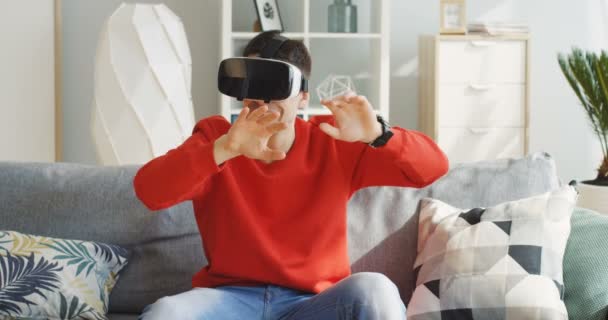 Portrait shot of the young caucasian man wearing a red sweater having a VR headset in the VR glasses on the couch with pillows in the nice light room. Indoors - Materiał filmowy, wideo