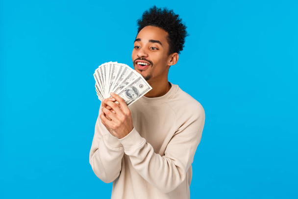 Finance, addiction and people concept. Excited and happy african american guy love money, express affection and admiration, kissing cash, counting dollars, smiling pleased, blue background - Photo, Image