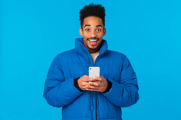 Excité heureux souriant afro-américain dans une veste d'hiver rembourrée, tenant smartphone et sourire optimiste, recevoir une fête d'invitation, bavarder avec des amis, est devenu célèbre sur les médias sociaux, fond bleu - Photo, image