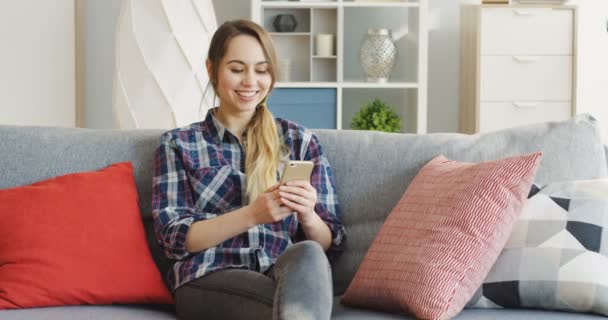 Jovem mulher bonita na camisa variegada sentado no sofá com travesseiros e conversando enquanto mensagens de texto no telefone inteligente. Para dentro
 - Filmagem, Vídeo