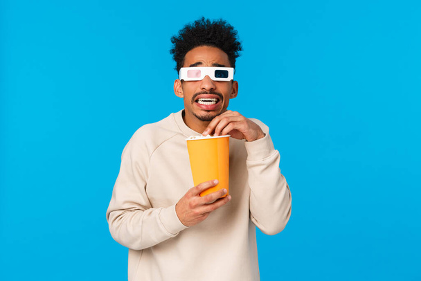 So scary. Trembling and terrified african-american hipster guy watching horror movie, frightening scene in film 3d glasses, holding paper box and eating popcron, standing tensed, blue background - Fotografie, Obrázek