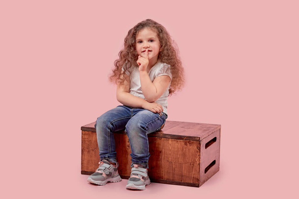 Beautiful little girl sitting on a wooden pedestal shows a gesture of silence. - Фото, изображение