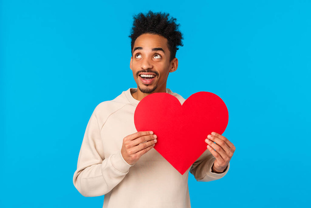 Drea,y and passionate, upbeat african american guy thinking how make perfect valentines day date, looking upper left corner thoughtful, holding heart card, searching true love, blue background - Фото, зображення