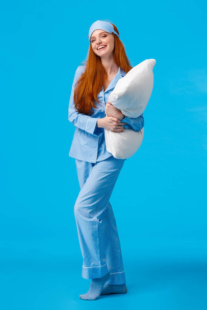 Full-length vertical studio cheerful lovely caucasian woman with ginger red long hair in nightwear, pyjama and sleep mask, hugging pillow ready sleep going bed at nigh and laughing, smiling - Фото, изображение