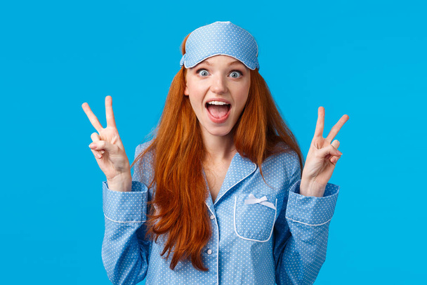 Aufgeregt nettes rothaariges Mädchen in Schlafmaske und Pyjama, das Friedenszeichen zeigt und schreit überwältigt positive Emotionen, steht amüsiert über blauem Hintergrund in Nachtwäsche - Foto, Bild