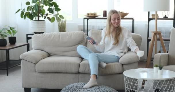 Frau benutzt Handy und trinkt Kaffee zu Hause - Filmmaterial, Video