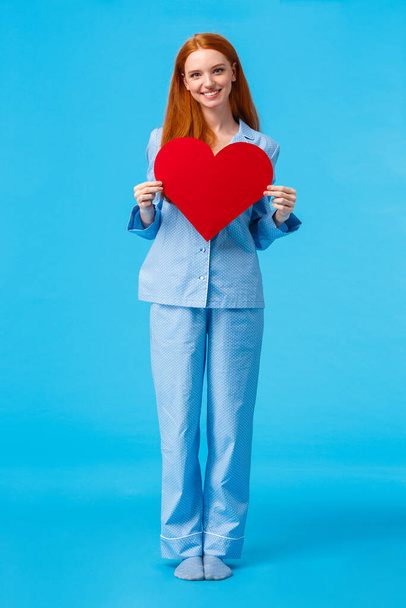 Vertical full-length portrait redhead elegant woman in cute pyjama, holding big red heart sign, valentines day gift and smiling, express affection, showing true feelings, confess, blue background - Photo, Image