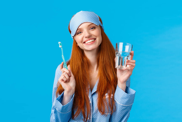 Happy smiling and carefree redhead enthusiastic girl in sleep mask, pyjama, holding glass water and toothbrush, taking care mouth hygiene, dental daily routine, blue background - Photo, Image