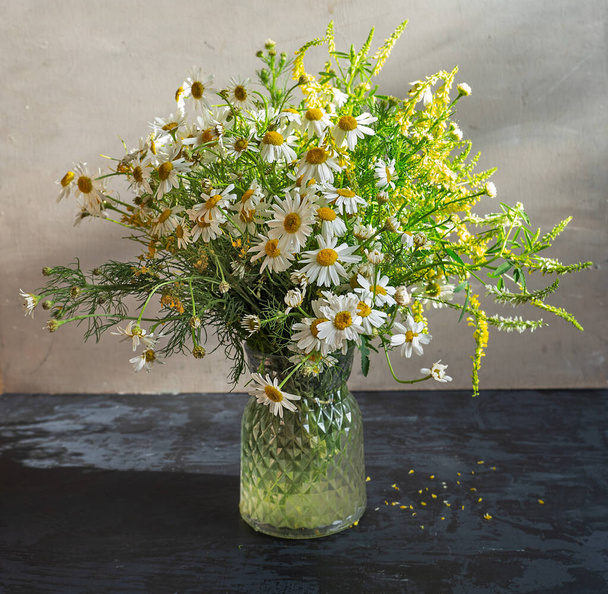 Licht boeket van wilde bloemen met de prevalentie van madeliefjes in een glazen vaas op een donkere tafel - Foto, afbeelding