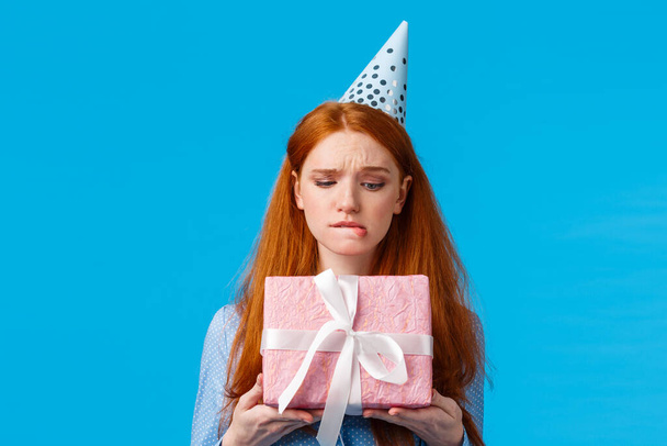 Заманчивый момент разворачивания подарка. Милая гламурная рыжеволосая девушка-подросток, кусающая губы, готовая открыть милый подарок на день рождения, носящая глупую кепку, празднующая день рождения стоящего голубого фона
 - Фото, изображение