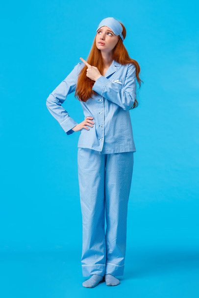 Uneasy and worried, concerned upset redhead female in nightwear and sleep mask, looking pointing upper left corner sad and distressed facing hard choice, standing troubled blue background - Фото, изображение