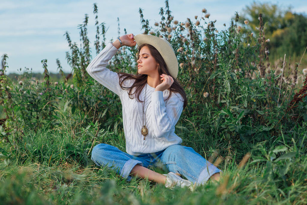 Beautiful brunette teenage girl dressed in a white sweater, jeans and cowboy straw hat sitting on green grass, smiling and laughing during sunset - Фото, изображение