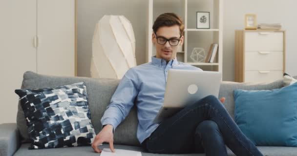 Young handsome businessman in glasses having a video chat on the laptop computer and checking the documents in his hand at home on the sofa. Indoors - Séquence, vidéo